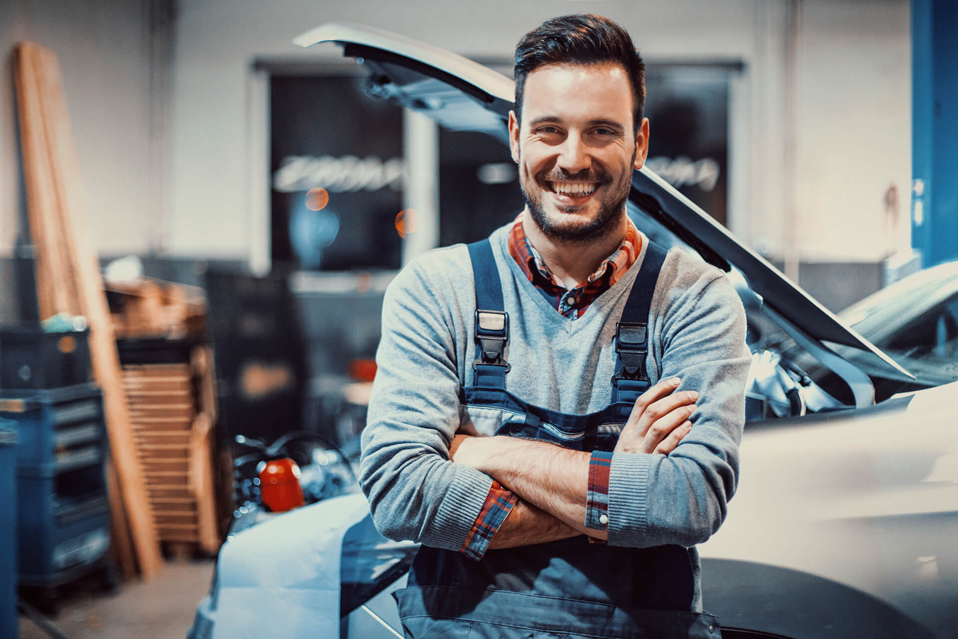 Mechaniker einer Autowerkstatt lehnt am Kotflügel eines defekten Fahrzeuges mit offener Motorhaube